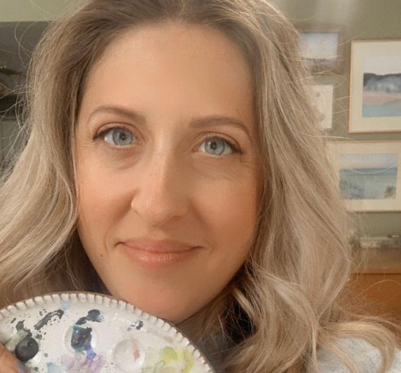 A woman with long blonde wavy hair and blue eyes holding a paint palette, smiling at the camera inside a room with framed pictures on the wall.