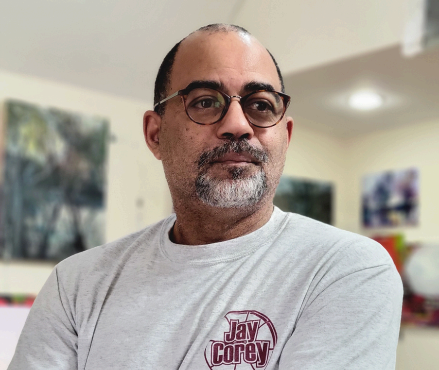 A man with glasses and a beard wearing a white T-shirt with the name 'Jay Corey' on it. He is standing indoors with artwork on the wall behind him.