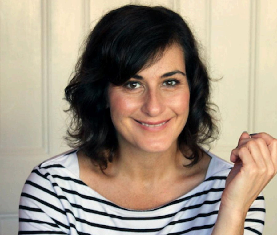 A woman smiling and making a fist gesture, with dark shoulder-length hair, wearing a striped white and black shirt, standing in front of a beige wall.