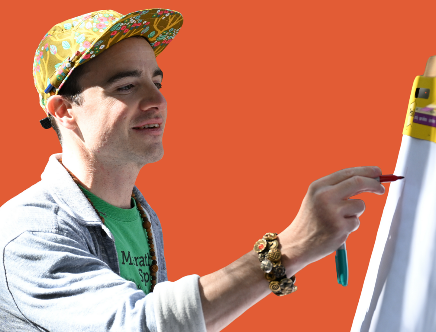 A man wearing a colorful cap, green shirt, and gray jacket is drawing on a whiteboard with a red marker in front of an orange background.