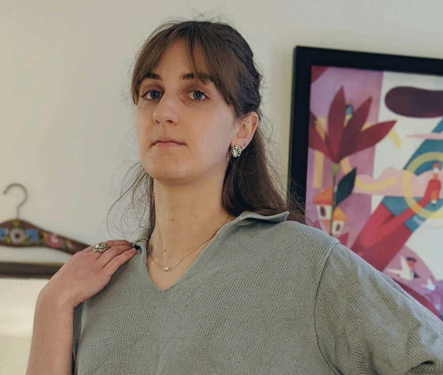 A woman with brown hair and blue eyes, wearing a gray sweater and jewelry, stands in front of an abstract colorful artwork on the wall.