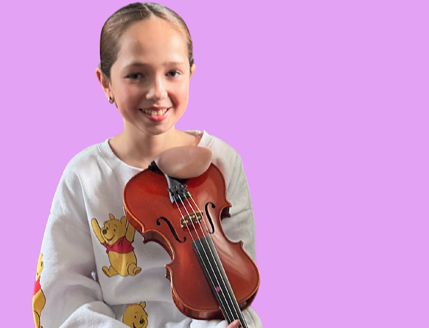 A young girl smiling and holding a violin against a purple background. She is wearing a white sweatshirt with Winnie the Pooh characters on it.