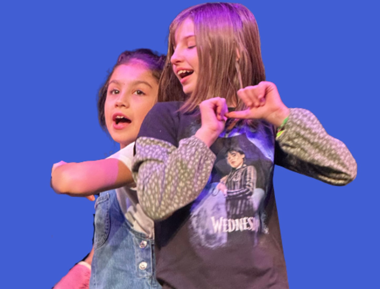 Two young girls, one with short hair and the other with long hair, stand against a blue background. The girl with long hair is wearing a black shirt with a graphic print, while the other girl is wearing a denim jacket. Both girls appear to be singing or performing.