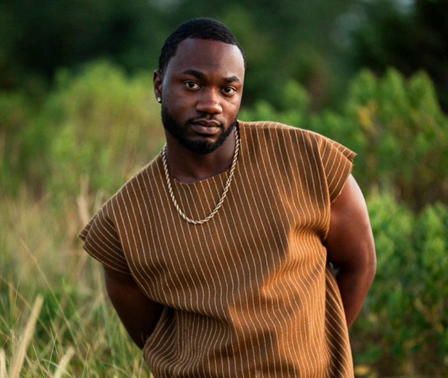 A man with dark skin, short black hair, and a beard poses outdoors in front of green foliage. He wears a brown striped sleeveless shirt, a gold chain, and earrings.