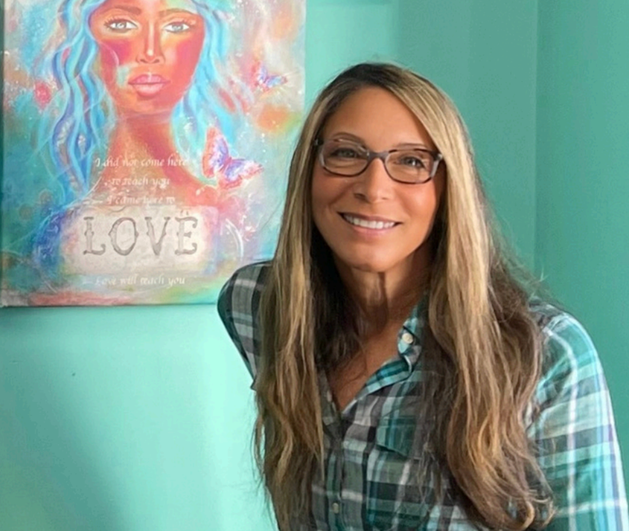 A woman with long wavy hair, wearing glasses and a plaid shirt, smiling at the camera, standing in front of a colorful painting of a woman with blue and pink hair and the word 'LOVE' on it.