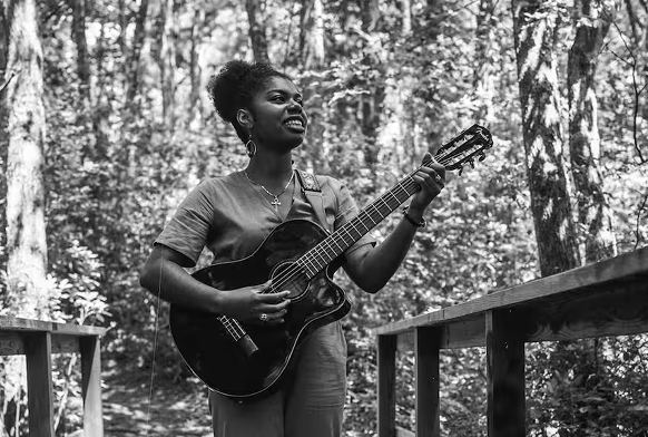A woman playing an acoustic guitar outdoors in a forest, smiling.