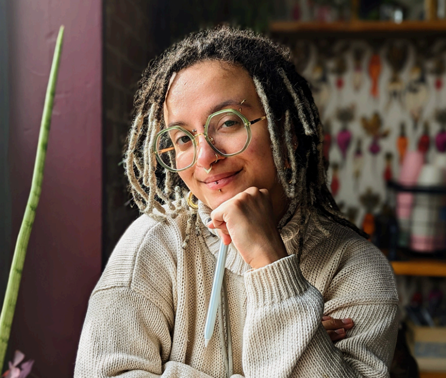 A woman with glasses and dreadlocks smiling, resting her chin on her hand, in an indoor setting.