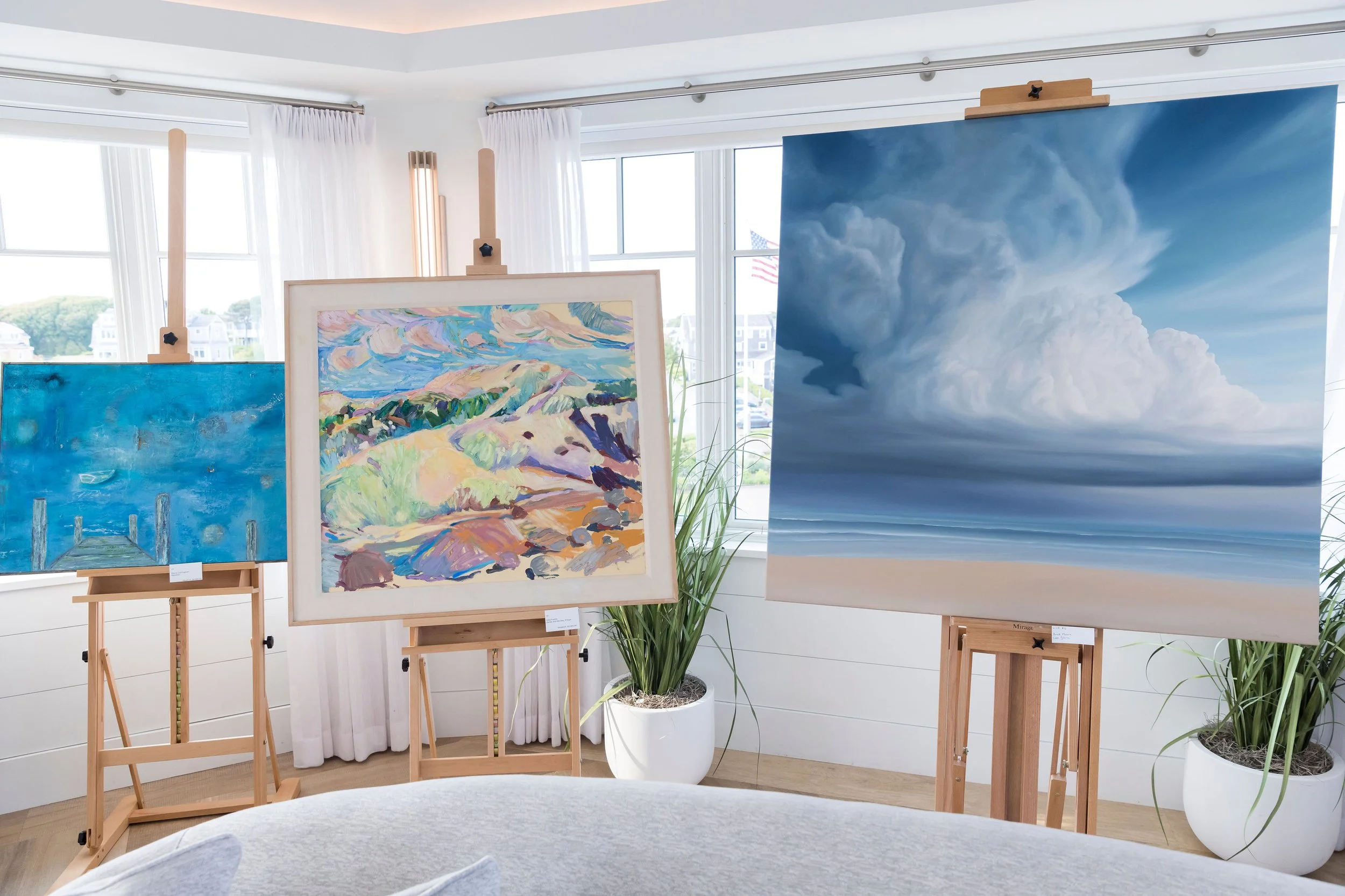 Arts Gala, Arts Foundation of Cape Cod, An art gallery displaying three landscape paintings, with two paintings on wooden easels and one on a stand in a bright room with white walls, windows, and potted plants.