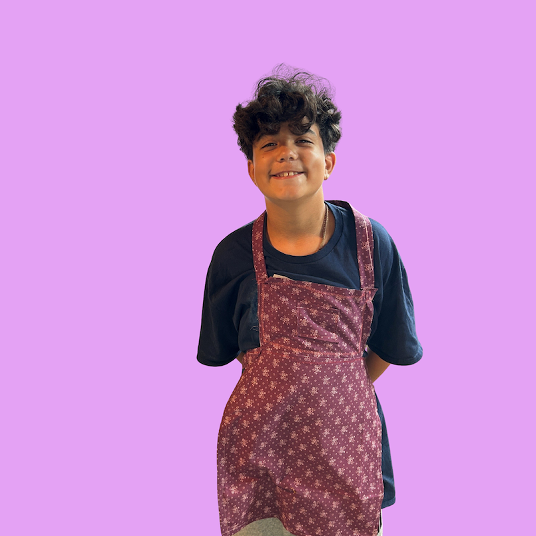 A smiling boy with curly dark hair wearing a black shirt and a maroon apron with white floral patterns, standing against a purple background.