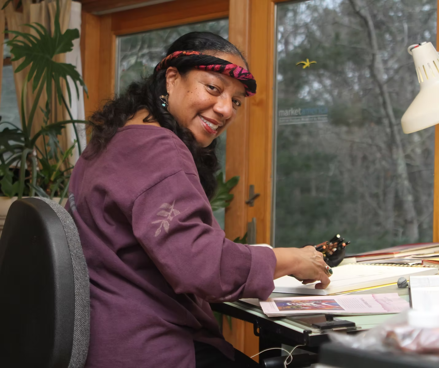 A woman with dark curly hair, wearing a purple sweatshirt and colorful headband, smiling at the camera while sitting at a desk in a room with large windows and greenery outside.
