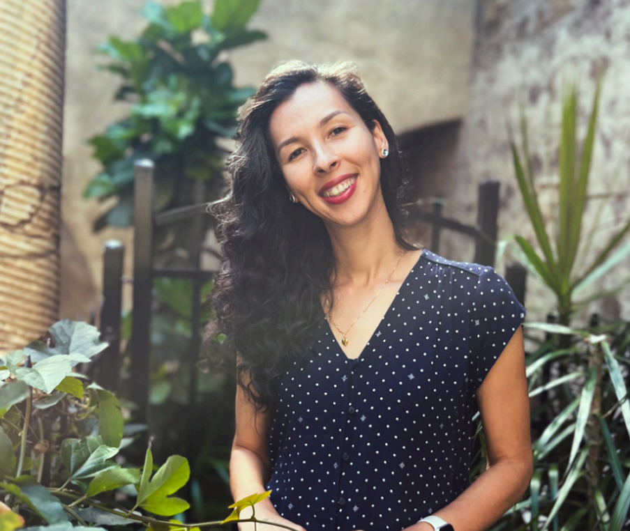 A woman with long, dark, curly hair, smiling, standing outdoors with green plants and a brick wall in the background.