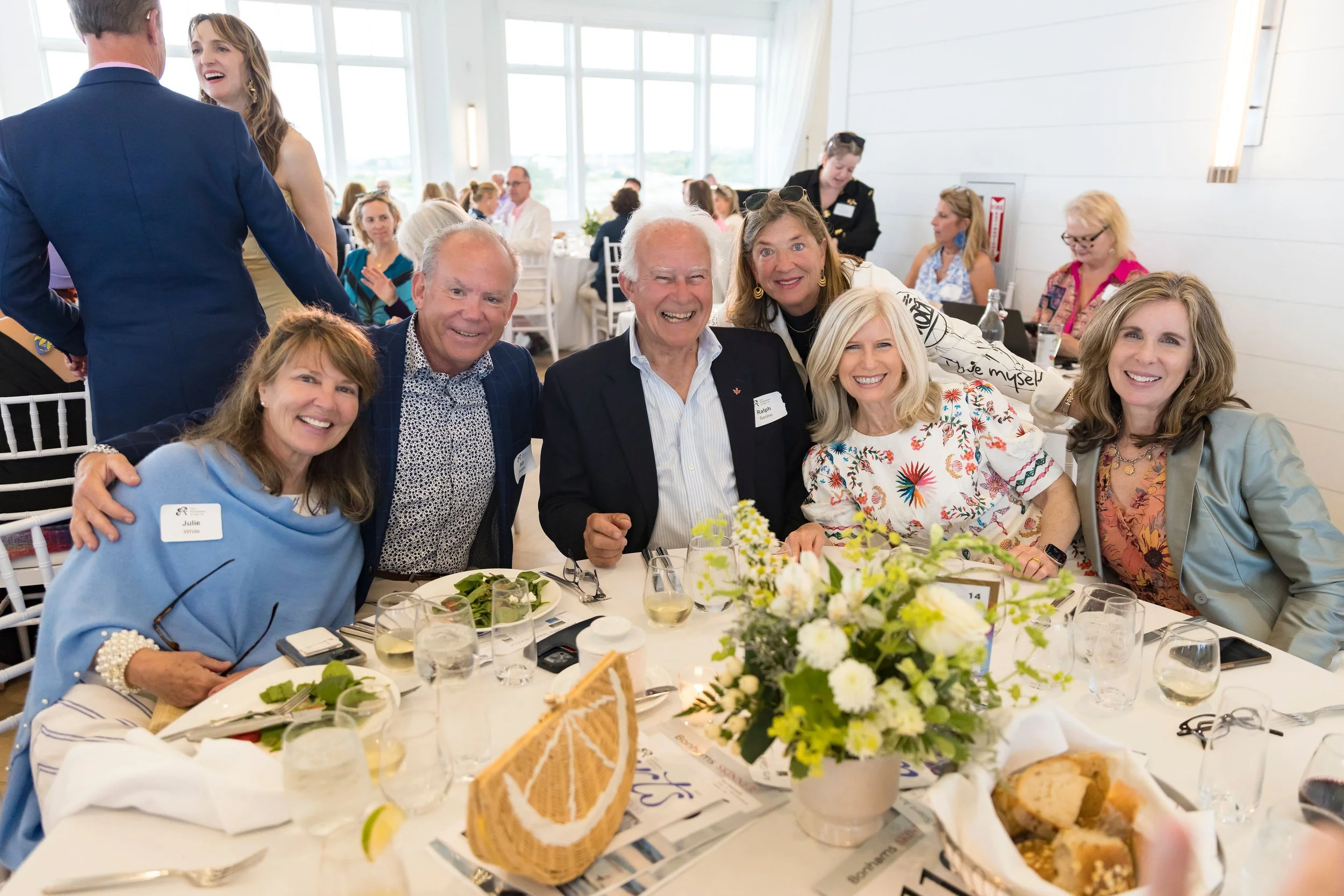 Arts Gala, Arts Foundation of Cape Cod, A group of six people at a banquet table, smiling for the camera, with glasses of wine and a flower arrangement, in a bright, modern venue.