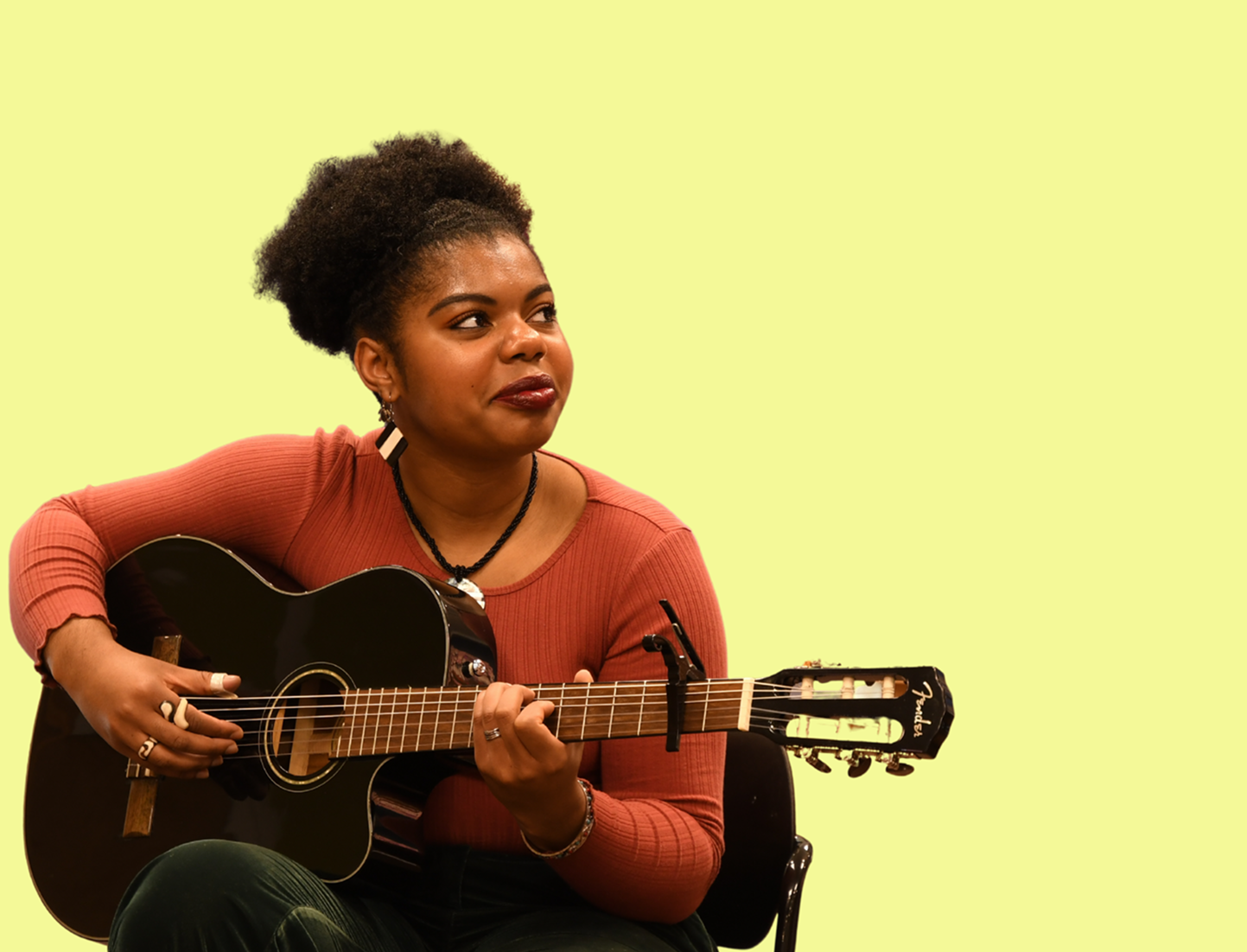 A woman with natural curly hair playing a black acoustic guitar, sitting in a chair against a light yellow background.
