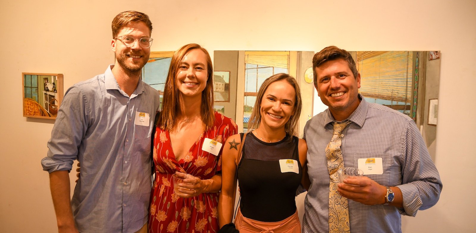 Four people at a social gathering smiling and holding drinks, standing in front of a colorful mural.