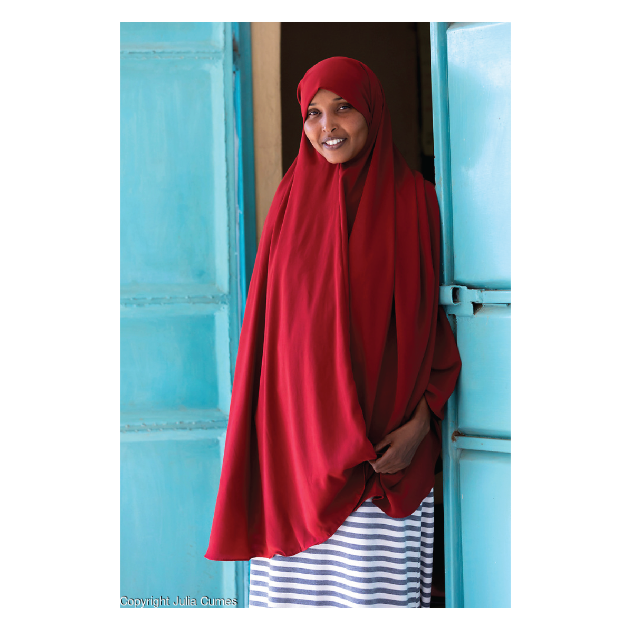 Julia Cumes Photography, Smiling woman wearing a red hijab and striped skirt standing in a doorway with light blue walls.