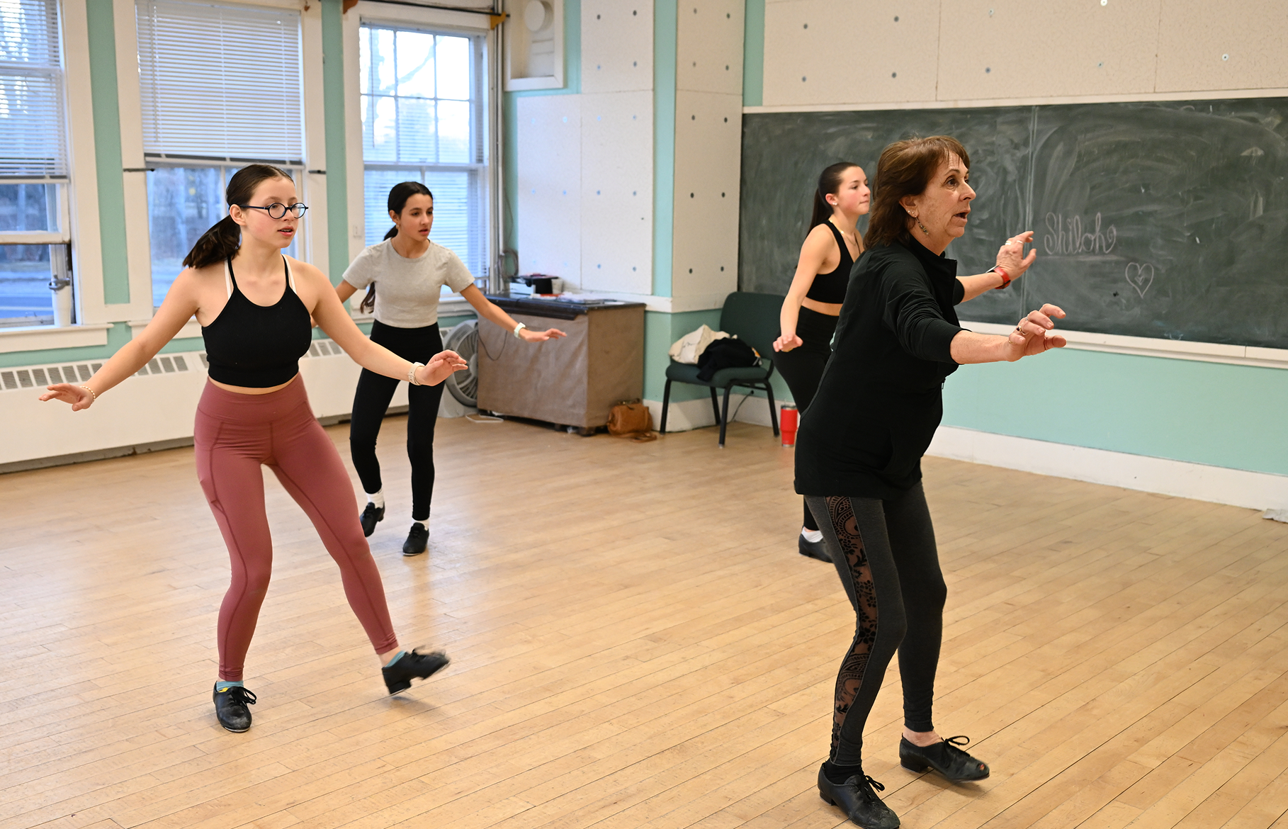 Four women participating in a dance or exercise class in a room with wooden flooring and large windows, with a blackboard on the wall.