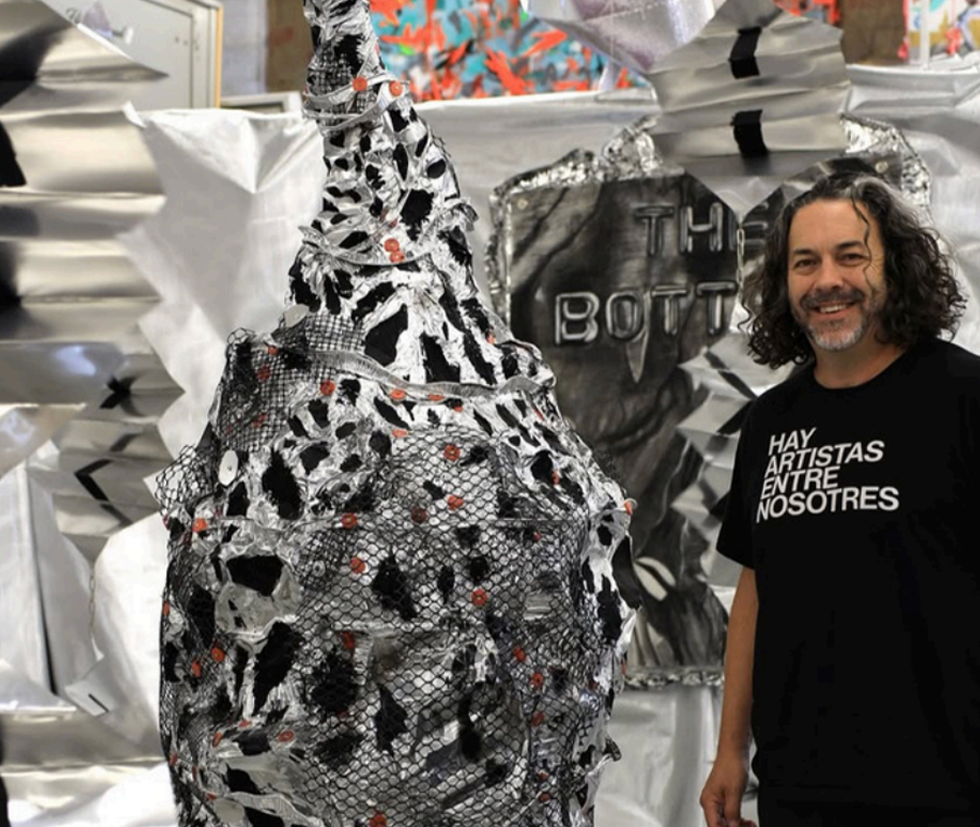 A man with curly hair and a beard wearing a black t-shirt that says "Hay Artistas Entre Nosotros" standing next to an abstract, black-and-white sculptural piece with red accents, inside an art studio or gallery with various artworks and materials in the background.