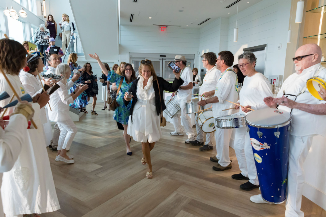 Arts Foundation of Cape Cod Gala, A woman in a black jacket and white dress dancing and smiling at a lively event with a band in white outfits playing drums and percussion, while other guests watch and take photos in a bright, modern indoor space.