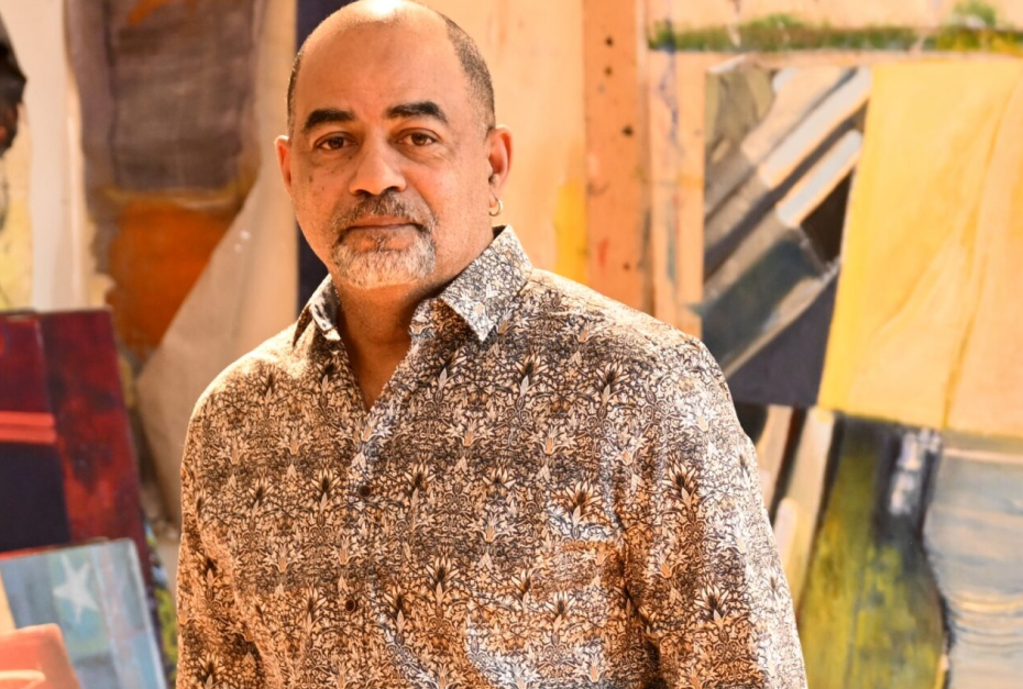 A middle-aged man with a beard and bald head, wearing a patterned shirt, standing in front of colorful abstract paintings in an art gallery or studio.