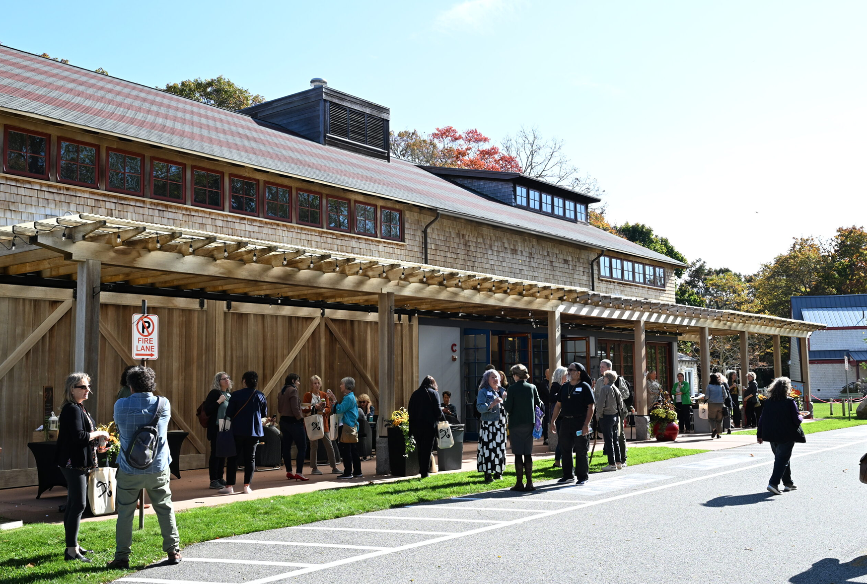People standing in line outside a rustic building with a wooden facade, under a sunlit sky, with trees in the background.