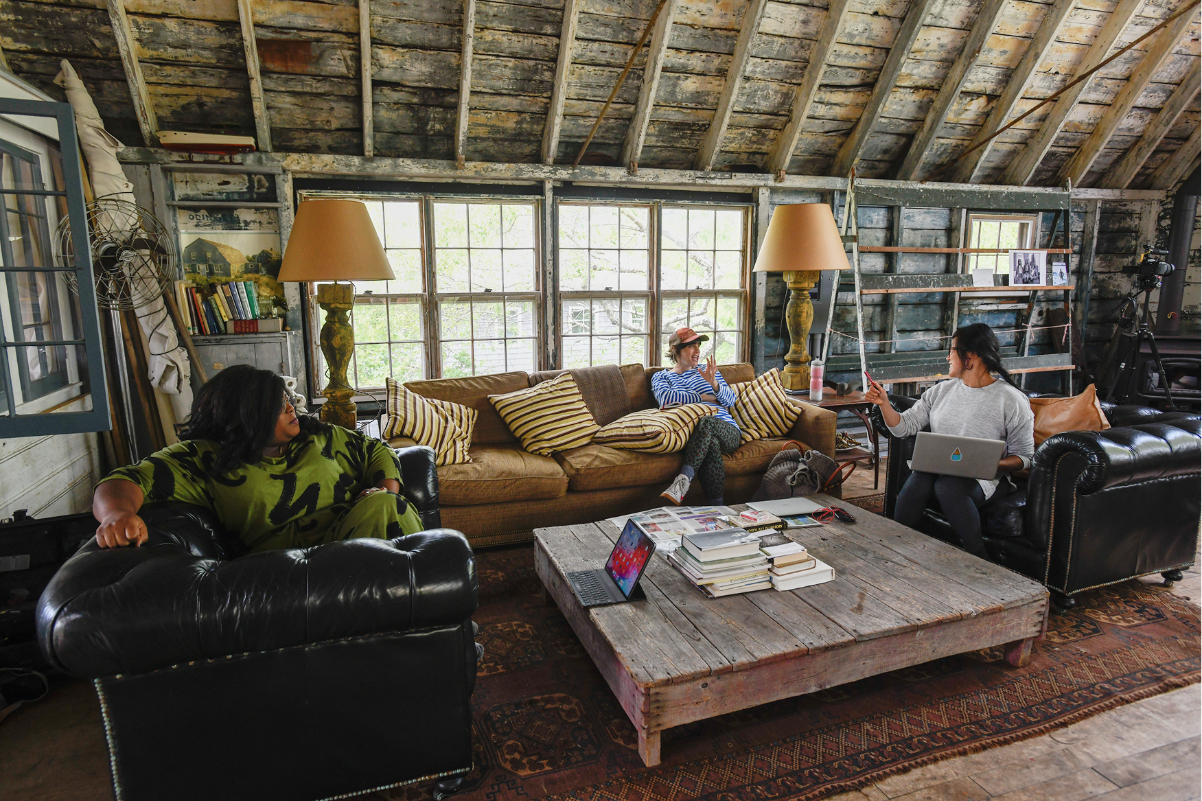A rustic living room with three women, two on black leather chairs and one on a tan couch, in a cozy, cluttered setting with large windows, bookshelves, and a wooden coffee table with books and a laptop.