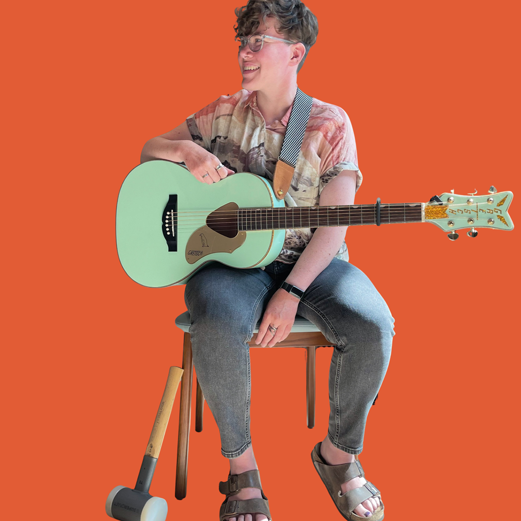Person sitting on a chair holding a light green acoustic guitar, wearing casual clothing, with an orange background.