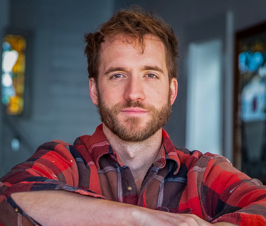 A man with light skin, brown hair and a beard, wearing a red and black plaid shirt, sitting with his arms crossed, with a blurred background in what appears to be a cozy indoor setting.