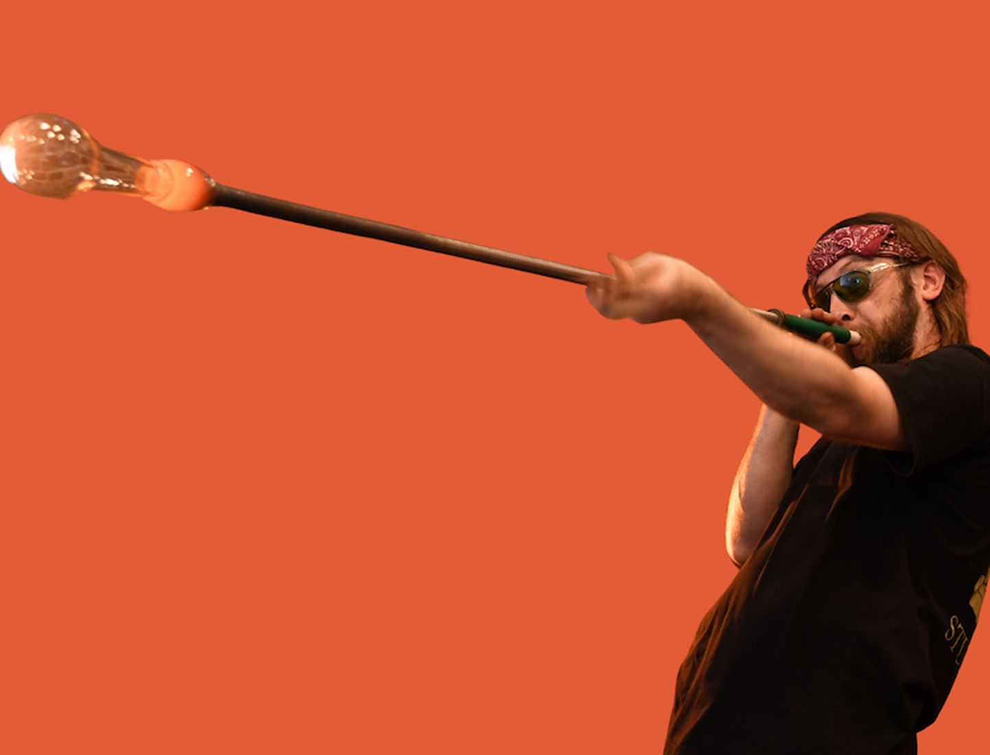 A man with a beard, bandana, and sunglasses playing a didgeridoo against a solid orange background.