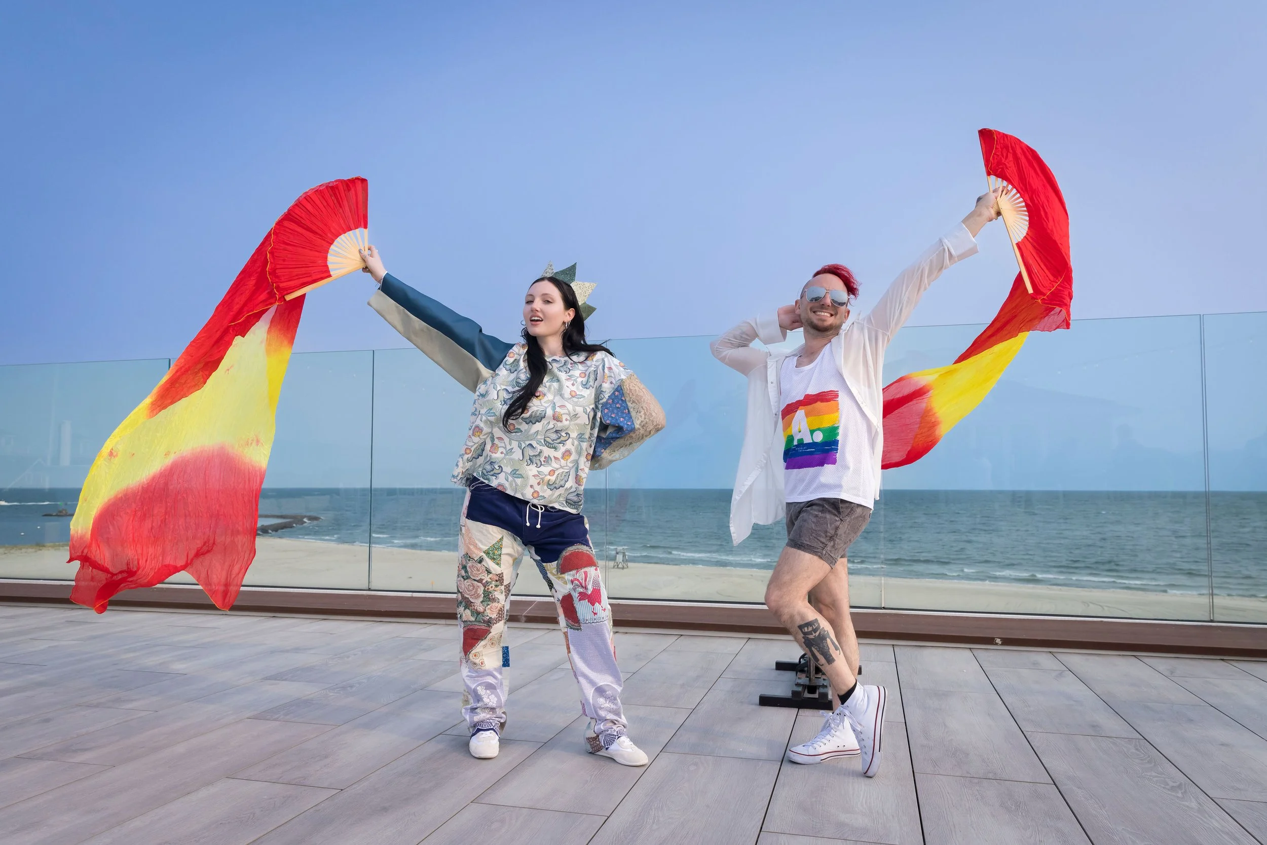 Arts Gala, Arts Foundation of Cape Cod, A couple holding rainbow-colored fans at the beach, smiling and dressed casually, with a glass barrier and an ocean in the background.