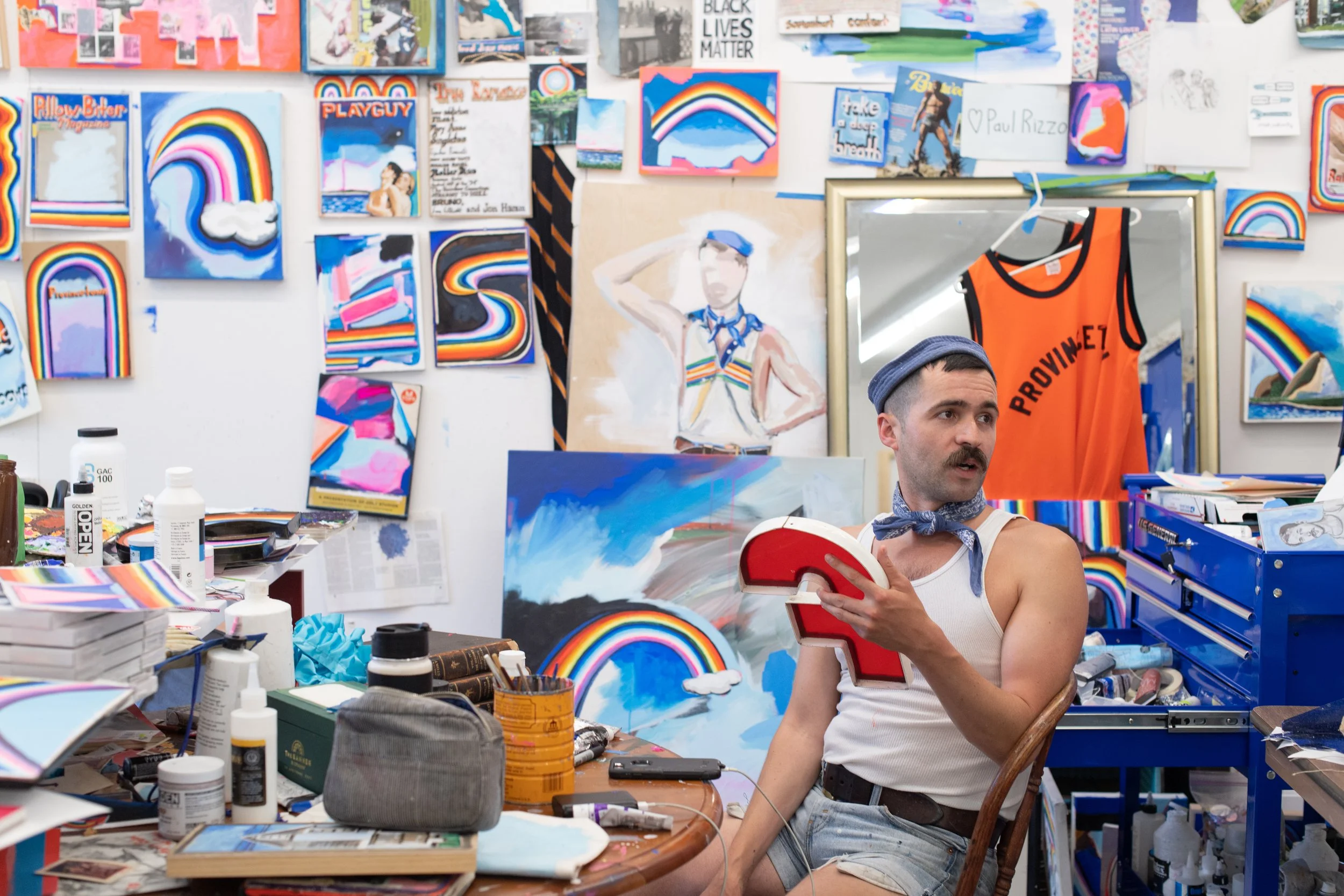 A man with a mustache wearing a blue cap, white tank top, and a blue bandana around his neck sits in an art studio. He holds a red question mark prop and looks to the side. The cluttered studio has colorful art pieces, paintings of rainbows, and a mannequin painted with a rainbow design. The wall is filled with various artworks, and there is a blue tool chest next to him.