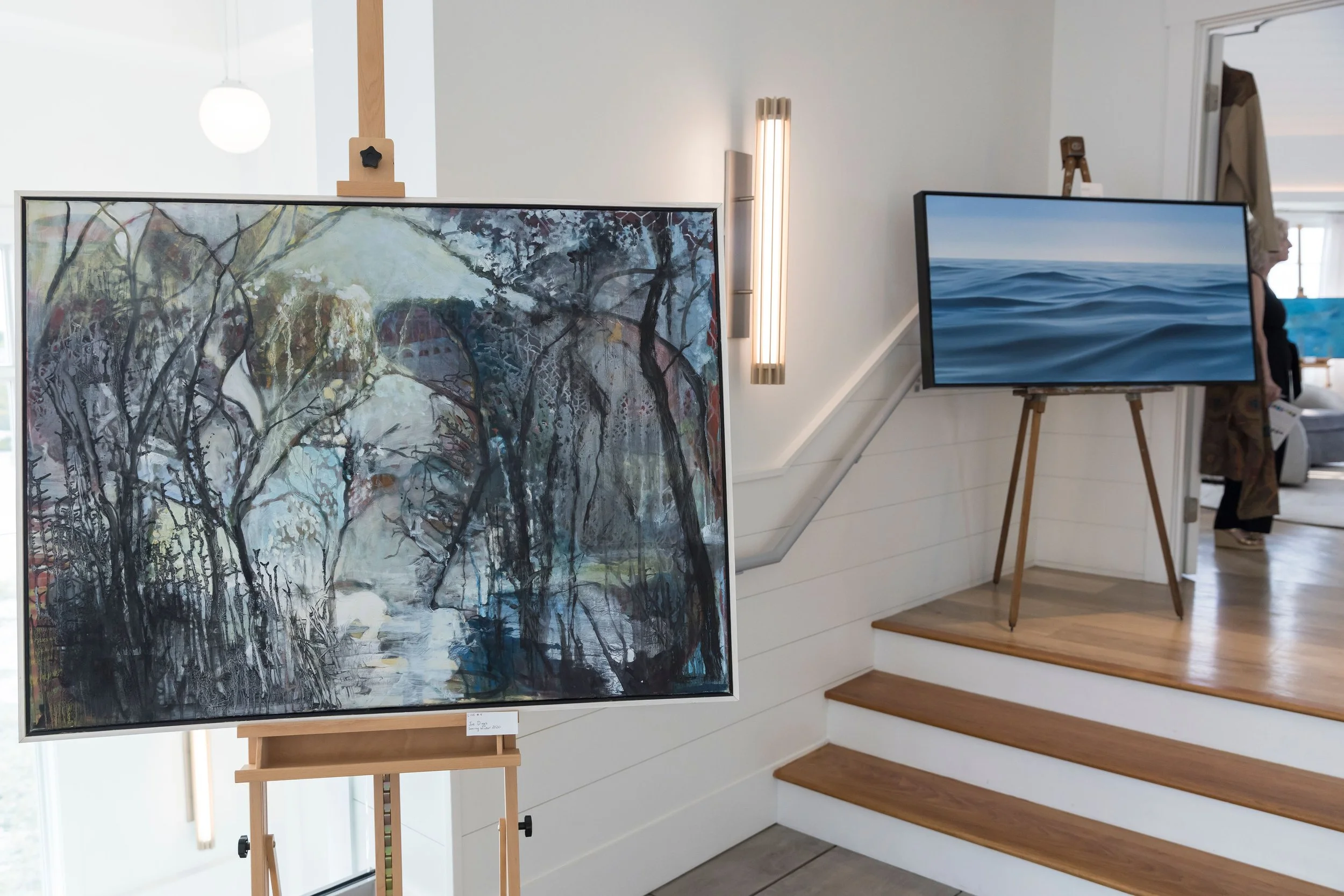 Arts Gala, Arts Foundation of Cape Cod, Art gallery display with a painting of a landscape featuring trees and a river on an easel, and a digital screen showing an ocean scene, set near wooden stairs and white walls.