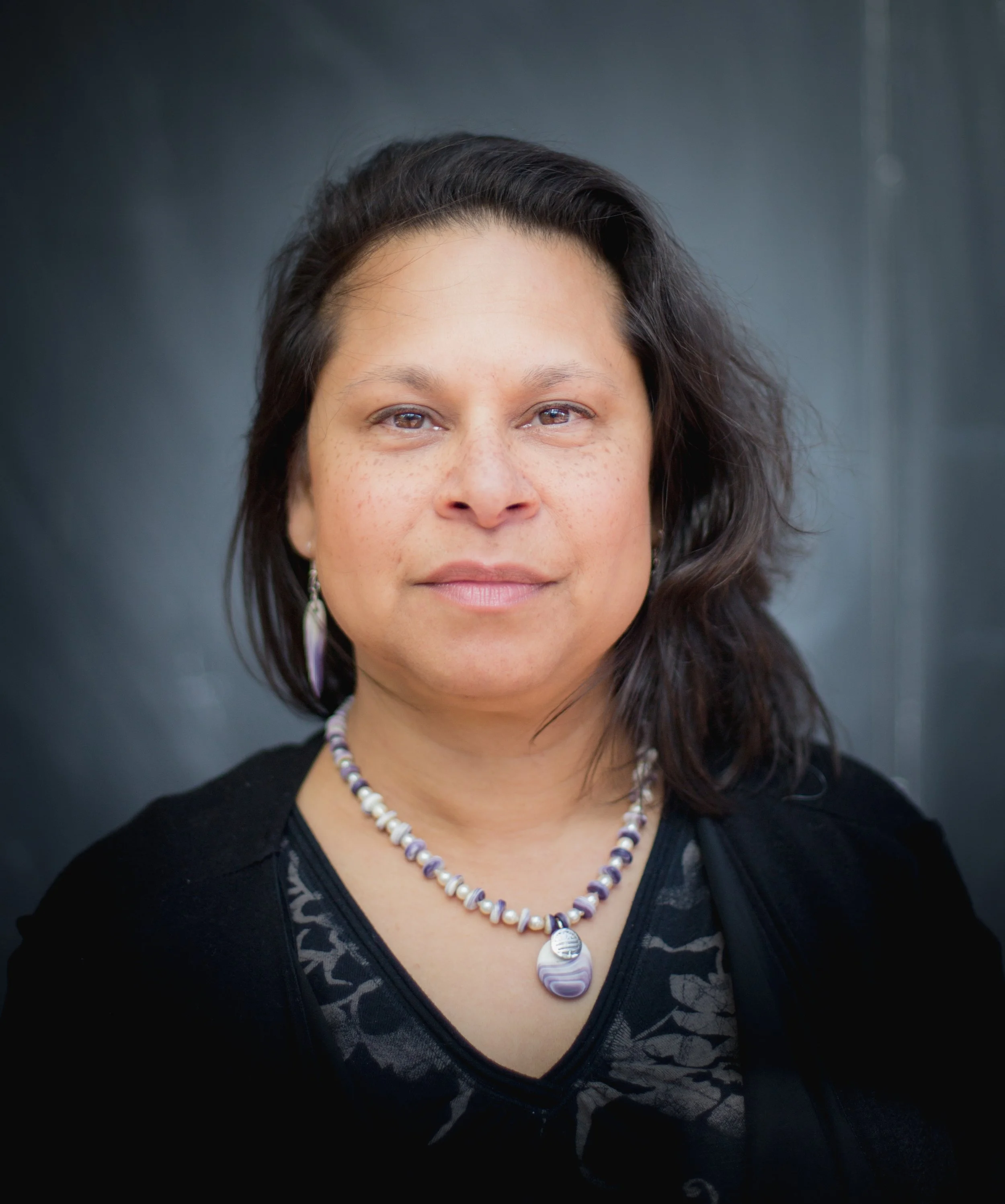 A woman with shoulder-length dark hair wearing earrings and a beaded necklace, looking directly at the camera with a neutral expression.