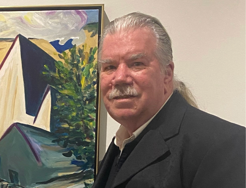 A man with long gray hair, mustache, and light complexion wearing a black coat standing next to a colorful painting of buildings and trees on a wall.