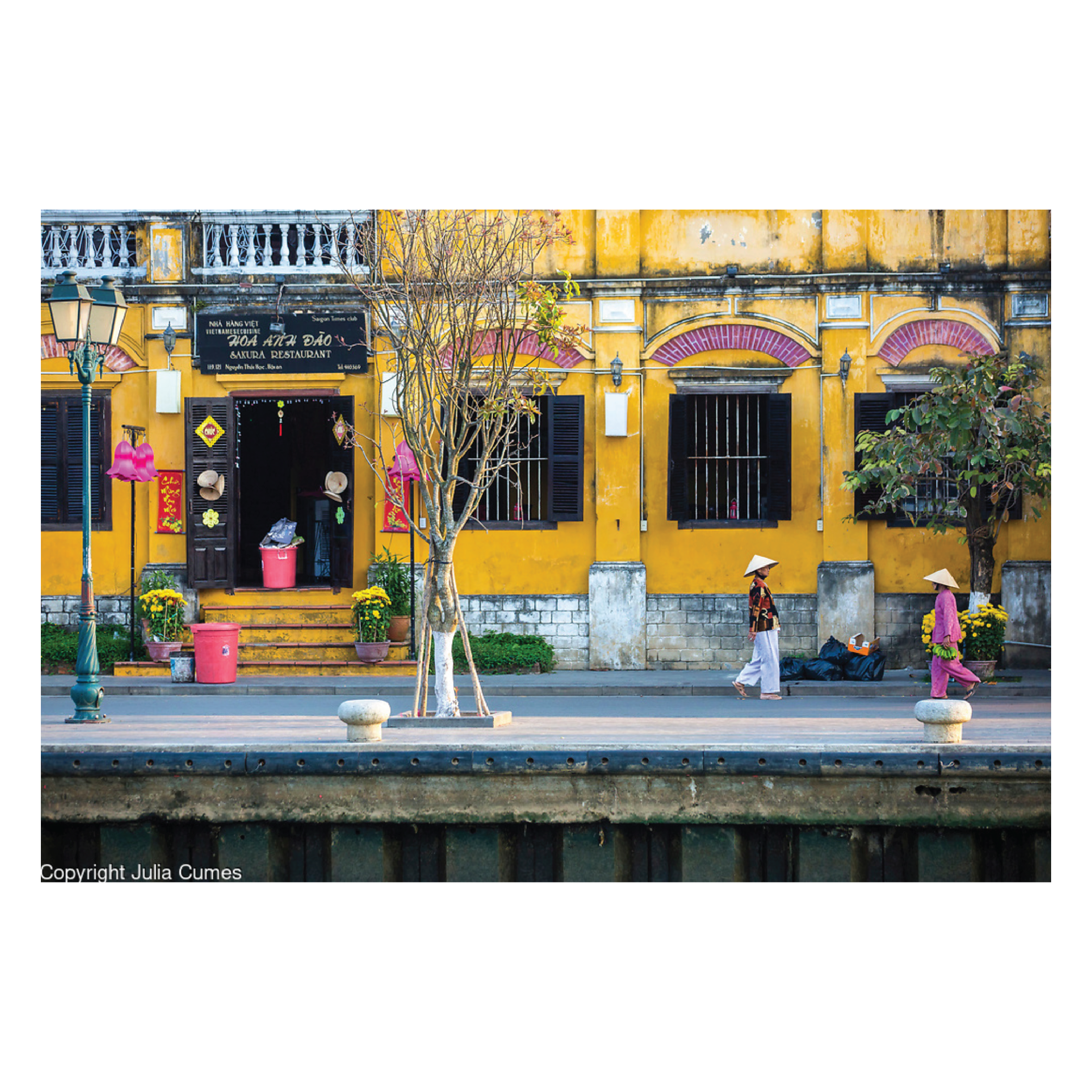 Julia Cumes Photography, Vietnamese street scene with a yellow building, two women in traditional conical hats walking on the sidewalk, and trees with yellow flowers.