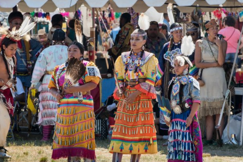Indigenous women and girls dressed in colorful traditional regalia at a cultural festival known as a powwow, with other attendees and vendor booths in the background.