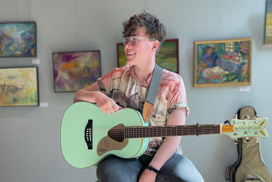 A person with short curly hair, glasses, and a patterned shirt sitting and holding a light green acoustic guitar while smiling, with colorful paintings on the wall behind them at an art gallery.