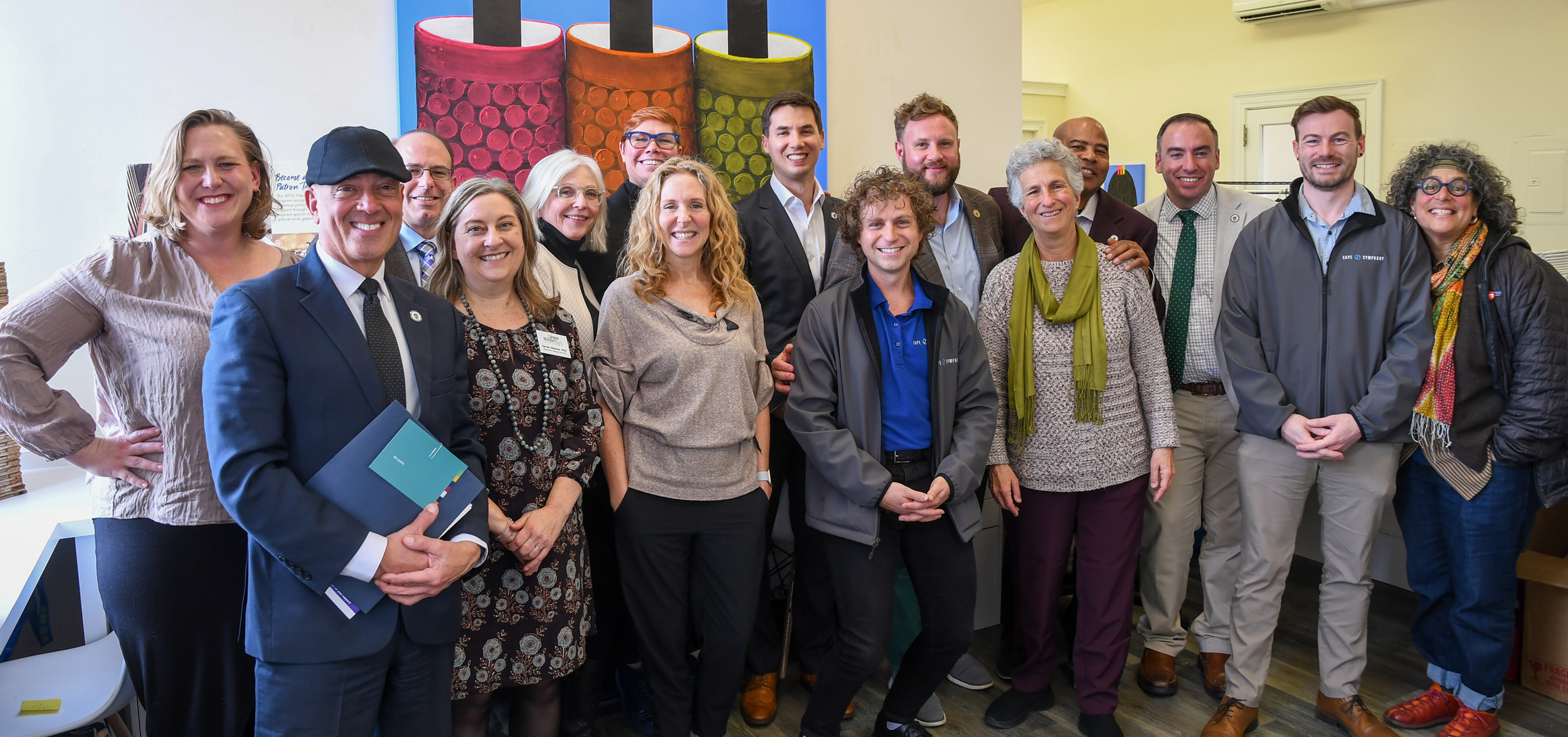 A group of 15 diverse people standing together indoors, smiling for the camera, with colorful artwork on the wall behind them.