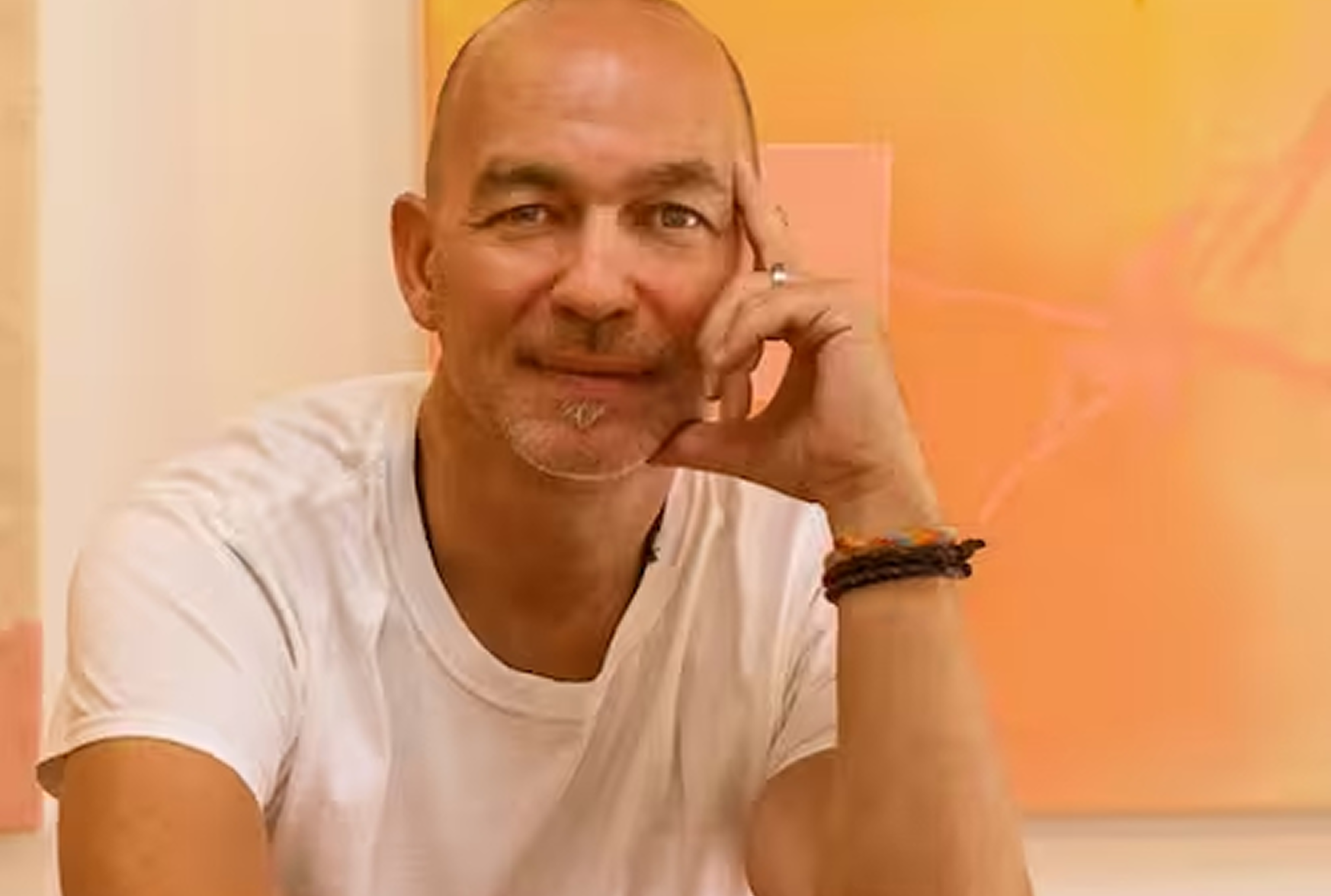 A middle-aged man with a shaved head and goatee, sitting at a table, resting his chin on his hand, smiling at the camera. He is wearing a white T-shirt and a black bracelet.