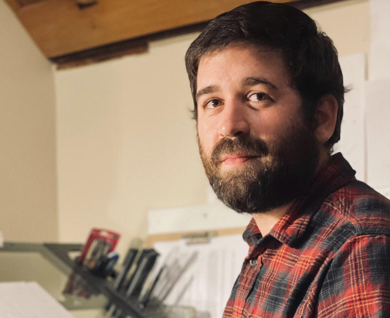 A man with dark hair and a beard, wearing a red and black plaid shirt, looking at the camera in an indoor setting.