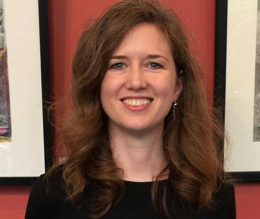 A woman with long, wavy red hair smiling at the camera in front of a red wall with framed pictures.