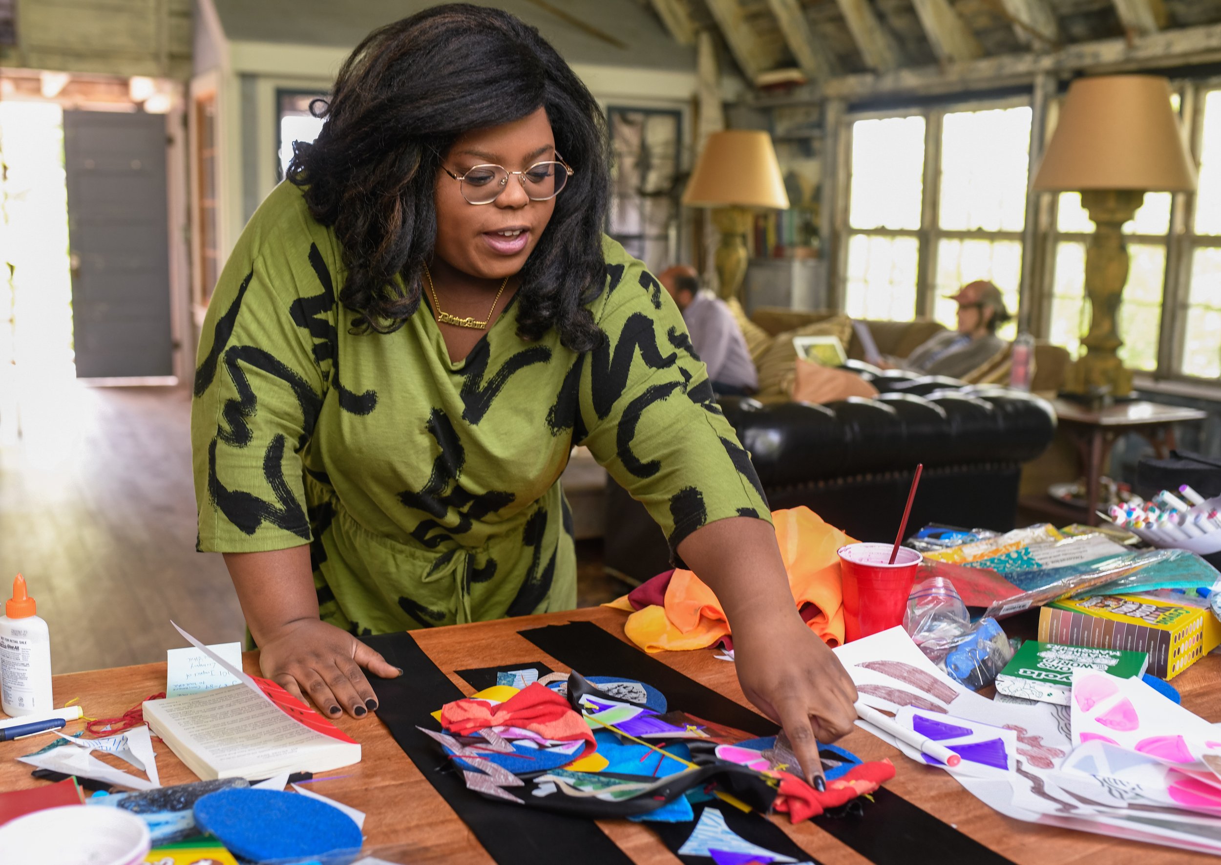 Woman in green and black patterned dress arranging fabric pieces on a table with art supplies, while two people sit on a couch in the background in a cozy, well-lit room.