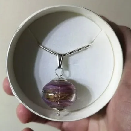 Hand holding a white circular box containing a necklace with a large, round purple and gold patterned pendant.