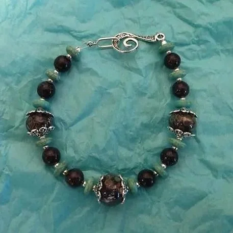A beaded bracelet with alternating dark purple and light green beads, featuring ornate silver accents and a decorative hook clasp, lying on crumpled teal-colored paper.