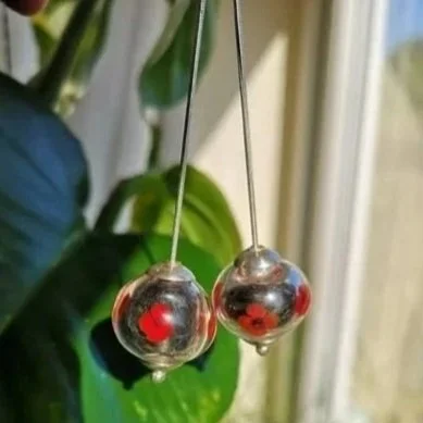 Silver earrings with red and black floral designs hanging in front of green leafy plants near a window.