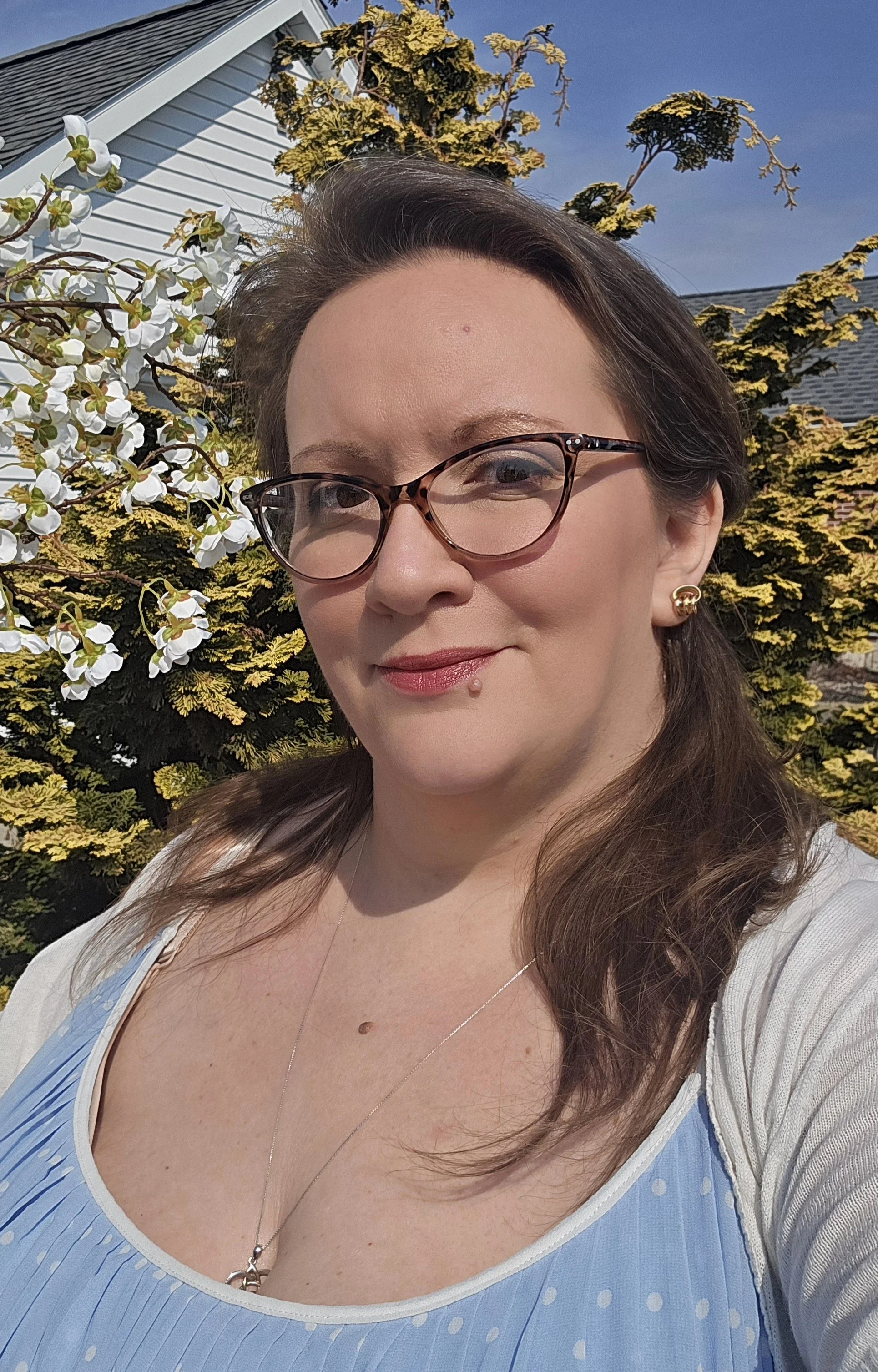 A woman with brown hair, glasses, and makeup taking a selfie outdoors. She is in front of a flowering shrub with white blossoms and yellow-green foliage, with a house and a blue sky in the background.