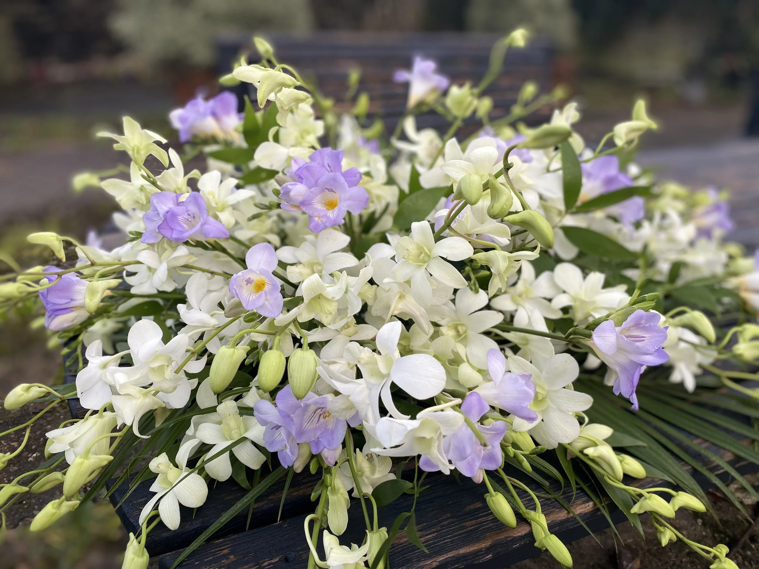 A bouquet of white and light purple flowers with green leaves, placed on a dark wooden bench outdoors.