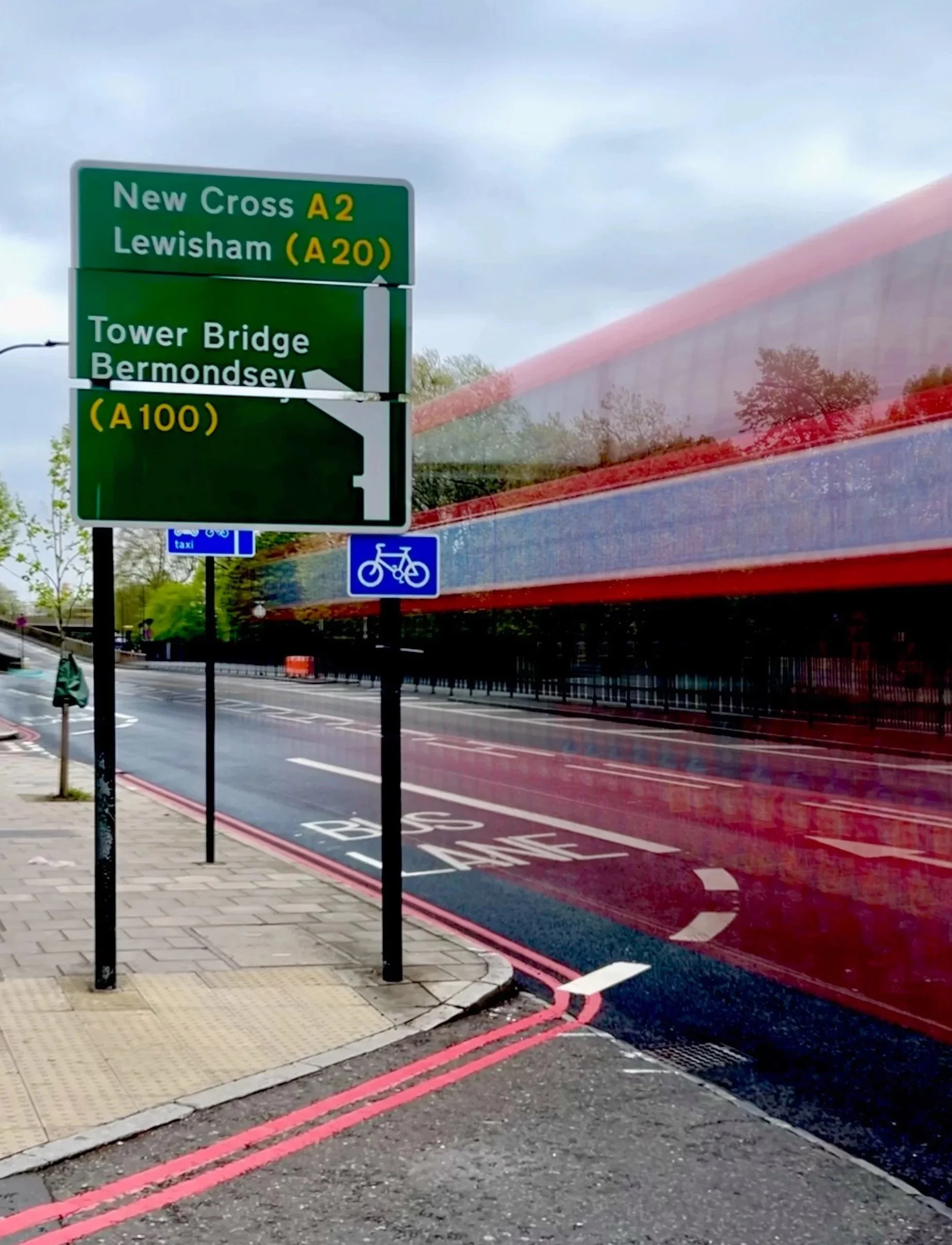 Green road signs with directions to New Cross, Lewisham, Tower Bridge, and Bermondsey on an urban street, with a blurred red double-decker bus passing by.