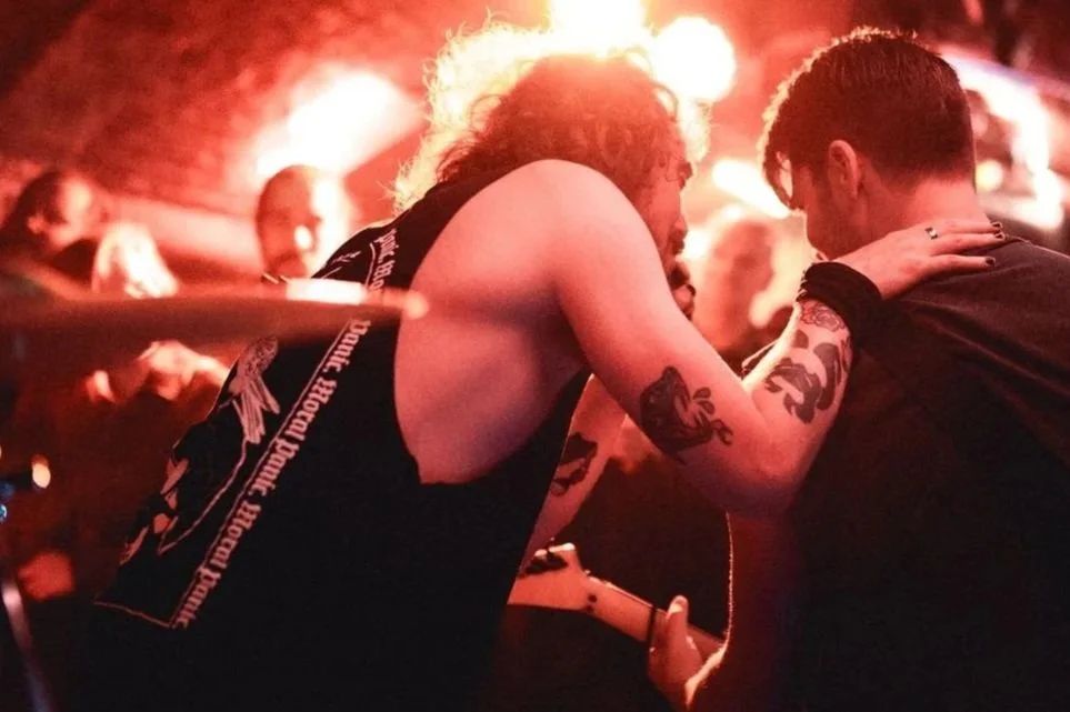 Two young men with tattoos leaning towards each other in a dimly lit, fiery environment, possibly a concert or club.