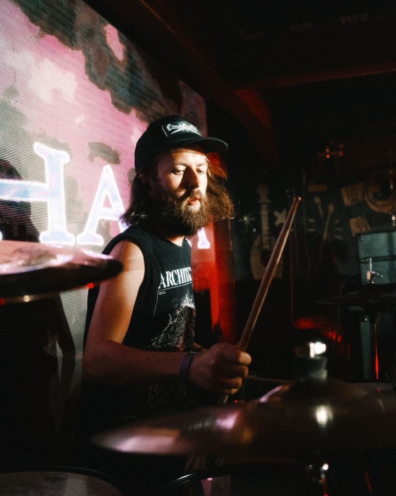 A musician with long hair, beard, wearing a black sleeveless shirt and cap, playing drums in a dimly lit venue with a pink and black background and neon lights.
