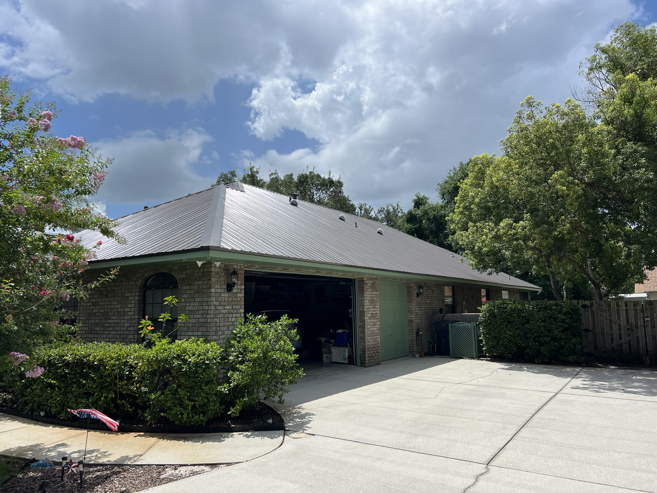 A residential house with an open garage, brick walls, a gray metal roof, and surrounding greenery including trees and bushes. The driveway is paved, and the sky is partly cloudy.
